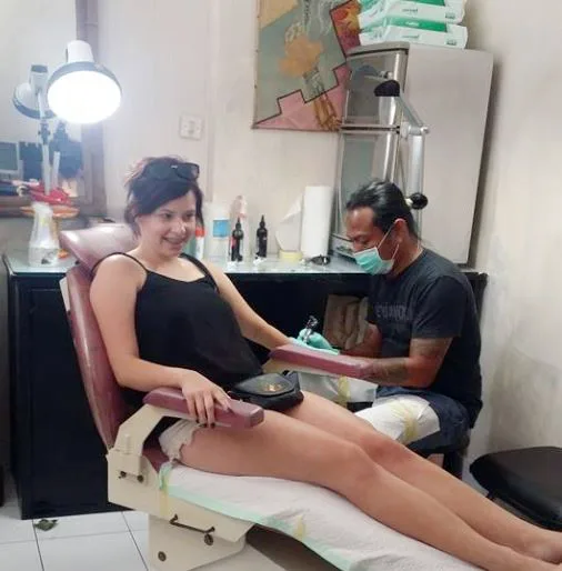 Masked artist tattooing a smiling client's leg